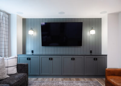 Omaha Home, Kitchen, Bathroom and Basement Remodeling - A modern living room featuring a wall-mounted flat-screen tv above a stylish grey cabinet, flanked by two wall sconces, with a cozy grey sofa and a leather armchair on the sides.
